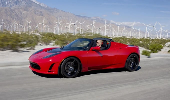Ein roter Tesla Roadster auf einer Landstraße zwischen Windrädern